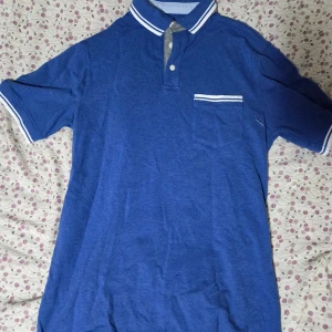 Blå henley från Merona med vit detalj - Blå henley från Merona i storlek S, med vit randig detalj på krage och ärmslut samt en bröstficka med vit rand. Skjortan har korta ärmar och knappar vid halsen. Perfekt för dig som gillar enkel och stilren design.