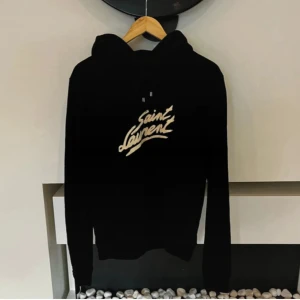 Saint Laurent - Svart hoodie från Saint Laurent med ikoniskt vitt logotyptryck på bröstet. Klassisk huva med snörning och långa ärmar. Tillverkad i mjuk bomullsmix för en skön och avslappnad vibe.