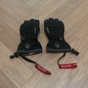 Svarta Hestra vinterhandskar - Säljer ett par svarta Hestra handskar med lång mudd och justerbart band runt handleden. Handskarna har röda säkerhetsremmar med Hestra-logga och är perfekta för kalla dagar. Materialet är slitstark syntet och designen är stilren med diskreta detaljer.