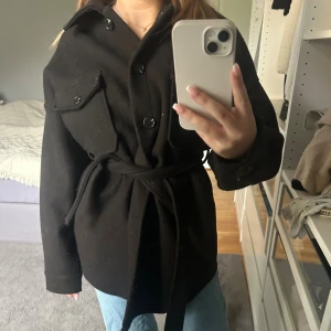 Svart kappa med knappar - Säljer en svart kappa med knappar och bälte runt midjan från Nelly.