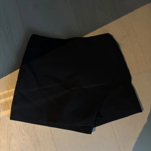 Svart omlottkjol från Zara - Svart kort omlottkjol från Zara med stilren design och två dekorativa fickor bak. Kjolen har en rak passform och är tillverkad i ett slätt tyg som ger en snygg och clean look. Perfekt för dig som gillar minimalistisk stil.