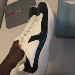 Prada sneakers i svart och vitt - Snygga Prada sneakers med vit mesh och svarta lackdetaljer. Skorna har rund tå, platt sula och klassisk snörning. Röd logga på plösen ger en exklusiv touch. Perfekt för dig som gillar stilrena och lyxiga sneakers med modern vibe.