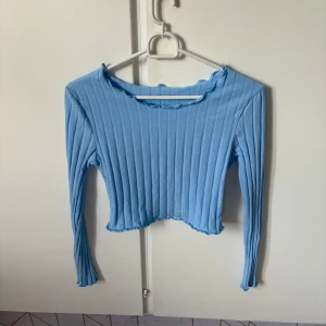 Ljusblå ribbad topp från SHEIN - Säljer en ljusblå långärmad topp från SHEIN med ribbad struktur och croppad passform. Toppen har en lätt vågig kant vid halsringning och nederkant, vilket ger en extra söt detalj. Perfekt för dig som gillar stilrena och trendiga plagg.