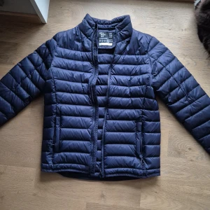 Mörkblå jacka från Neomondo - Snygg mörkblå jacka från Neomondo i juniorstorlek 14 år. Jackan har quiltad design, hög krage och dragkedja framtill. Tillverkad i syntetmaterial som håller dig varm och bekväm. Perfekt för kyliga dagar och enkel att matcha med din stil.