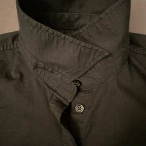 Svart skjorta Ted Bernhardtz strl 48 - Snygg svart skjorta från Ted Bernhardtz med korta ärmar och klassisk krage. Skjortan har en figurnära passform och knappar hela vägen framtill. Materialet är mjukt och känns som bomull, perfekt för en clean och stilren look.