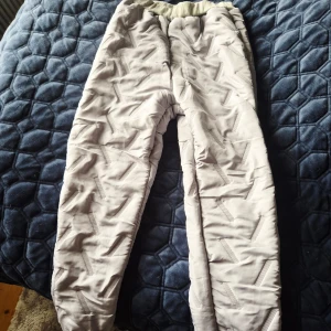 Gråa överdragsbyxor med mjukt foder - Säljer ett par gråa täckbyxor med snyggt mönster och mjukt, fluffigt foder på insidan. Byxorna har resår i midjan för skön passform och är perfekta för kalla dagar. De har ett quiltat yttre och detaljer i form av diskreta meshpaneler.