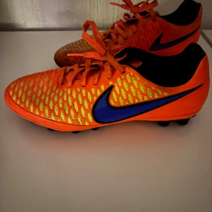 Nike Magista orange fotbollsskor - Nike Magista fotbollsskor i starkt orange med blå swoosh och gul mönstrad ovandel. Skorna har snörning, rund tå och platt sula med fasta dobbar för gräsplan. Ovandelen är tillverkad i syntetmaterial och insidan är svart. Perfekt för dig som vill sticka ut på planen.
