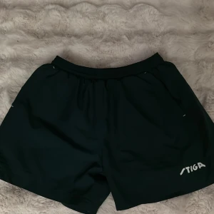 Svarta träningsshorts från Stiga - Mörkblåa shorts från Stiga i storlek small, perfekta för träning. De har elastisk midja och är tillverkade i ett lätt och skönt syntetmaterial. Klassisk loose passform och enkel design med Stiga-logga på benet. Snygga att ha på gymmet eller till sport.