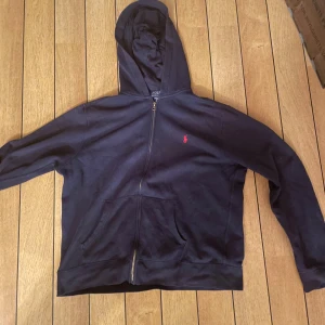 Mörkblå hoodie från Polo Ralph Lauren - Snygg mörkblå hoodie från Polo Ralph Lauren med dragkedja och huva. Klassisk röd logga på bröstet, två fickor framtill och mjukt bomullsmaterial. Perfekt för chill dagar och enkel att matcha med jeans eller mjukisbyxor. Det står Xl men då den är väldigt liten i storleken så är den ungefär Small till Medium