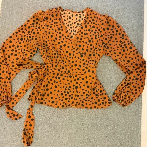 Omlott blus med leopardmönster - Säljer en snygg omlott blus i orange med svart leopardmönster. Blusen har långa puffärmar med resår vid ärmslut och knytband i midjan. V-ringad hals och lätt volang vid nederkant ger en trendig look. Perfekt för dig som gillar att sticka ut!