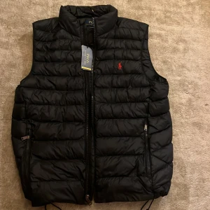 Mörk blå dunväst från Polo Ralph Lauren - Snygg mörk blå dunväst från Polo Ralph Lauren med röd broderad logga på bröstet. Västen har quiltad design, två dragkedjefickor och hög krage. Materialet är polyester och nylon, vilket gör den lätt och bekväm. Perfekt att slänga över en hoodie för extra värme. Har aldrig använd den,köpte den för 4600