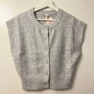 Ljusgrå stickad väst med knappar - Mysig ljusgrå stickad väst från Nelly med rund halsringning och knäppning framtill. Västen har bred ribbad kant nedtill och vid ärmöppningarna, vilket ger en avslappnad och trendig look. Perfekt att slänga över en skjorta