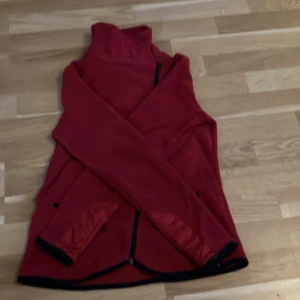 Röd fleecejacka med svarta detaljer storlek 151-160 - Röd fleecejacka med svart dragkedja och svarta detaljer vid fickorna och ärmsluten. Jackan har hög krage och två fickor med dragkedja framtill. Materialet är mjukt och värmande, perfekt för kyliga dagar. Långärmad och enkel, med stilren look.