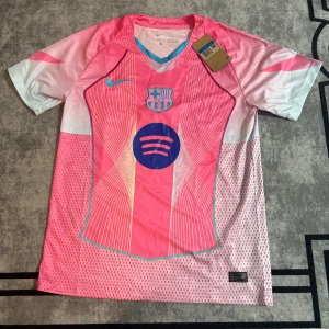 Barça x Nike rosa fotbollströja M - Snygg FC Barcelona fotbollströja från Nike i rosa och vitt med tunna linjer och mönster. Tröjan har klubbmärket och Spotify-loggan på bröstet, samt en V-ringad krage med blå detalj. Tillverkad i lätt, ventilerande Dri-FIT polyester. Perfekt för match eller träning.