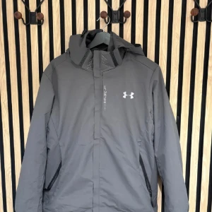 Grå vindjacka från Under Armour - Snygg grå vindjacka från Under Armour med huva och dragkedja. Jackan har två sidofickor med dragkedja och ett mjukt fleecefoder på insidan. Perfekt för blåsiga dagar och har en stilren look med logga på bröstet. Perfekt för skidor.