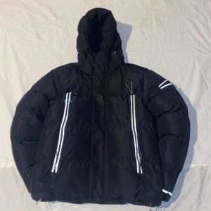 Svart pufferjacka från Lorenzo - Svart pufferjacka från Lorenzo med huva och reflekterande detaljer på dragkedjor och ärm. Jackan är vadderad, har två stora fickor framtill och är tillverkad i polyester. Perfekt för kalla dagar med sin isolerade och vindtäta design.