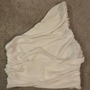 Vit oneshoulder topp i glansigt tyg - Säljer en vit oneshoulder topp med snygg drapering och elastisk kant nedtill. Tyget har en lätt glansig finish som ger en lyxig känsla. Perfekt för dig som vill sticka ut med en unik och trendig siluett.
