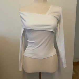 Vit offshoulder långärmad topp - Säljer en vit långärmad topp med offshoulder-design och asymmetrisk halsringning. Toppen är ribbad och har en tight passform som framhäver figuren. Perfekt för dig som gillar en stilren och modern look. Oanvänd 