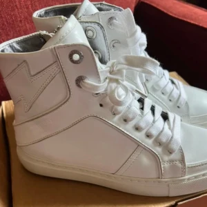 SÖKER  - Säljer ett par stilrena vita höga sneakers från Saint Laurent med snörning och cool blixtdetalj på sidan. Skorna är i skinn och har en klassisk platt sula. Perfekta för dig som gillar en clean och modern look.