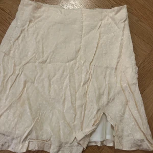 Beige kortkjol från Gina Tricot - Snygg beige kortkjol från Gina Tricot med subtilt leopardmönster i tyget. Kjolen har en liten slits framtill och är tillverkad i ett glansigt material som ger en lyxig känsla. Perfekt för dig som gillar stilrena och trendiga plagg.