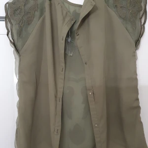 Blus - Olivgrön blus från Oxygene med transparenta partier och broderade detaljer i blommigt mönster. Blusen har tryckknappar framtill och en unik, vågig nederkant. Perfekt för dig som vill ha något annorlunda och stilrent i garderoben. Har ett hål.