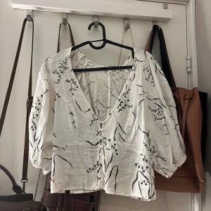 Vit blommig blus från H&M i linneblandning - Supersöt vit blus från H&M i linneblandning med svarta broderade blommor, v-ringning och knappar framtill. Blusen har puffiga långärmar och en luftig, lite croppad passform. 💕