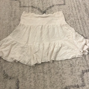 Vit volangkjol med smock i midjan - Supersöt vit kortkjol med bred smock i midjan och volang nertill. Kjolen har en luftig och flowy känsla, perfekt för varma dagar. Enkel att matcha med crop tops eller stickade tröjor.