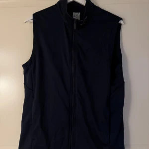 Mörkblå sportväst från Callaway - Snygg mörkblå sportväst från Callaway med hel dragkedja framtill och högt krage. Västen har tre vita pilar som detalj på ryggen och är tillverkad i ett lätt syntetmaterial som passar perfekt för träning eller golf. Enkel och stilren design.