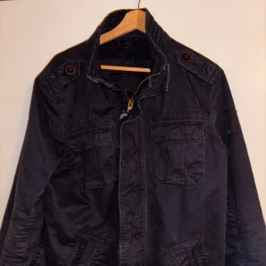 Mörkblå fieldjacket med fickor och dragkedja - Mörkblå fieldjacket med hög krage, flera fickor med lock och snygga bruna knappar på axlar och ärmslut. Jackan har dragkedja framtill och är tillverkad i bomullsmaterial. Perfekt för dig som gillar en clean och funktionell look.