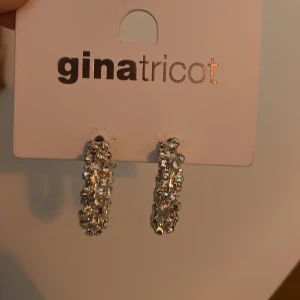 Glittriga silverörhängen från Gina Tricot - Säljer ett par snygga örhängen från Gina Tricot i silverfärg med glittriga stenar. De är halvcirkelformade och har en chunky design som verkligen drar blickarna till sig. Perfekta för att ge din look extra bling och att sticka ut i mängden.