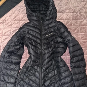 Svart dunjacka från Peak Performance - Säljer en svart dunjacka från Peak Performance med huva och snygga quiltade detaljer. Jackan har dragkedja framtill och Peak Performance-logga på bröstet. Perfekt för kalla dagar och har en modern, figurnära passform.