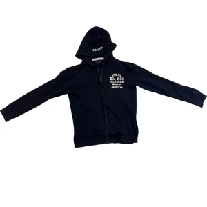 Svart hoodie från Tommy Hilfiger - Svart hoodie från Tommy Hilfiger med vit textprint på bröstet och huvan. Tröjan har dragkedja framtill, huva med tryck och två fickor. Perfekt för dig som gillar klassisk streetstyle och vill ha en snygg och enkel look.