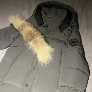 Grå dunjacka från Canada Goose - Säljer en grå dunjacka från Canada Goose med svart patch på ärmen och fluffig beige pälsdetalj på huvan. Jackan har knappar framtill och en hög krage som skyddar mot kyla. Perfekt för kalla vinterdagar och har en stilren look.
