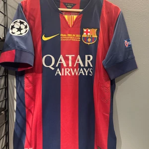 Barcelona Messi 10 Final Berlin 2015 tröja - Säljer en officiell FC Barcelona matchtröja från Final Berlin 2015 med Messi 10 på ryggen. Tröjan är randig i mörkblått och rött, har korta ärmar och är tillverkad i lätt, ventilerande polyester. Champions League-märke på ärmen och klubbmärke på bröstet. Helt ny med tagg en replika