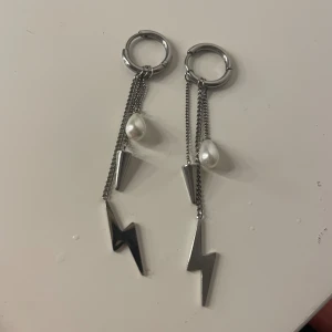 Örhängen med blixt och pärldetalj - Snygga örhängen i silverfärg med hängande kedjor, blixtformade hängen, konformad detalj och en vit pärla. Ger en cool och edgy vibe med mixen av metall och pärla. Perfekta för dig som vill sticka ut med accessoarerna.