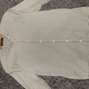 Grå skjorta från J.Lindeberg - Snygg grå skjorta från J.Lindeberg med klassisk krage och diskret broderad logga på bröstet. Skjortan har långa ärmar och knappar hela vägen framtill. Tillverkad i mjukt bomullstyg som känns skönt mot huden. Funkar t M också