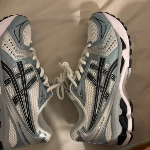 Asics Gel-Kayano 14 sneakers blå/vit - Säljer ett par Asics Gel-Kayano 14 sneakers i vitt, ljusblått och silver med svarta detaljer. Skorna har meshpaneler för extra andningsförmåga, klassisk snörning och en chunky sula med svart mönster undertill. Perfekta för dig som gillar sportig och trendig stil.