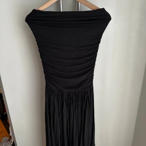 Svart bandeu långklänning från NA-KD - Svart långklänning från NA-KD med offshoulder-design och rynkad överdel. Kjolen är lätt veckad och klänningen har en elegant, figurnära passform upptill. Perfekt för dig som gillar stilrena och tidlösa plagg med en modern twist.