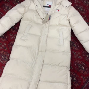 Beige lång dunjacka Tommy Jeans - Lång, beige dunjacka från Tommy Jeans med huva och dragkedja framtill. Jackan har quiltad design, två sidofickor och klassisk logga på bröstet. Perfekt för kalla dagar när du vill hålla stilen och värmen.