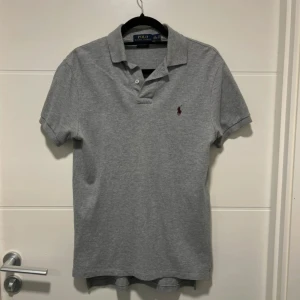 Grå pikétröja från Polo Ralph Lauren S - Snygg grå pikétröja från Polo Ralph Lauren i storlek S. Klassisk krage, två knappar och den ikoniska broderade loggan i vinrött på bröstet. Tröjan har korta ärmar och är tillverkad i mjuk bomull med en custom fit-passform.