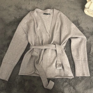 Grå stickad kofta med bälte - Snygg grå stickad kofta från Lager157 med omlott-design och matchande bälte i midjan. Koftan har långa ärmar med breda ribbade muddar och en mjuk, skön känsla. Perfekt för att hålla sig varm och ändå se chill ut.