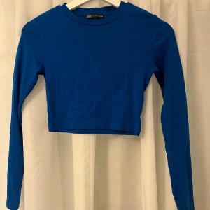 Blå croppad långärmad topp från Zara - Säljer en stilren blå croppad topp från Zara i storlek S. Toppen har lång ärm och rund halsringning, perfekt för att matcha med höga jeans eller kjol. Materialet är mjukt och stretchigt bomull som sitter skönt mot kroppen.