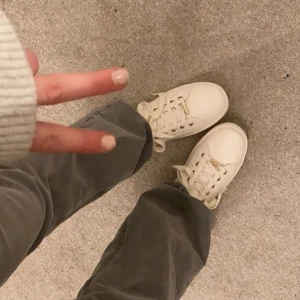 Vita sneakers med gulddetaljer - Snygga vita sneakers med guldfärgade detaljer vid hälen och snörhål. Skorna har rund tå, platt sula och snörning framtill. Materialet är skinn och de har dekorativa perforeringar längs sidorna. Perfekt för dig som gillar stilrena och trendiga sneakers.