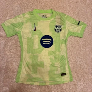 Barcelona Nike kortärmad fotbollströja - Barcelona fotbollströja från Nike i ljusgrön färg med geometriskt mönster. Tröjan har klubbmärket, Nike-logga och Spotify som sponsor framtill. UNHCR-logga på ryggen. Tillverkad i lätt och ventilerande polyester, perfekt för match eller träning.