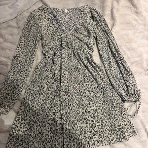 Vit blommig kortklänning med v-ringning - Supersöt vit kortklänning från H&M med svart blommigt mönster, v-ringad hals och långa ärmar. Klänningen har en lätt rynkad midja och flowig kjol, perfekt för vår och sommar. Tillverkad i mjuk polyester som känns skön mot huden.