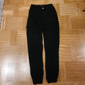 Svarta cargopants från Bik Bok, strl 34 - Svarta cargopants från Bik Bok med hög midja och elastiska muddar vid bensluten. Byxorna har stora fickor på sidorna och är tillverkade i ett mjukt bomullsmaterial. Perfekt för en cool och avslappnad streetstil.