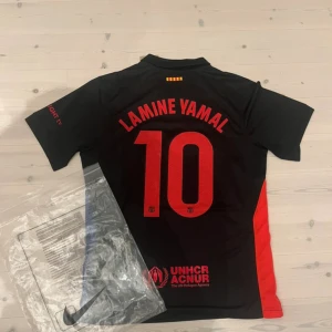 FC Barcelona x Cactus Jack Lamine Yamal tröja - Säljer en svart FC Barcelona x Cactus Jack fotbollströja från Nike med Lamine Yamal och nummer 10 på ryggen. Tröjan har röda och blå detaljer, korta ärmar och tryckta loggor samt UNHCR-märke. Materialet är lätt och andas, perfekt för match eller träning.