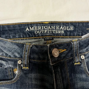 Blå capribyxor jeans  från American Eagle Outfitters - Snygga blå jeans i capri modell från American Eagle Outfitters med stretch och klassisk femficksdesign. Jeansen har en mörk tvätt med slitningar och kontrastsömmar, samt uppvikta benslut. Materialet är 98% bomull och 2% elastan för extra komfort.