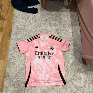 Rosa Real Madrid fotbollströja Adidas Medium - Säljer en snygg rosa Real Madrid fotbollströja från Adidas med svarta detaljer och tryck. Tröjan har korta ärmar, klubbmärke på bröstet och klassiska Adidas-ränder på axlarna. Materialet är lätt och andas, perfekt för träning eller match.