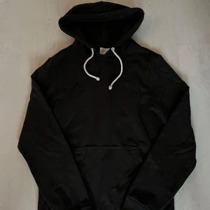 Svart hoodie från Pier One - Svart hoodie från Pier One med vita snören i huvan och klassisk känguruficka framtill. Tröjan har lång ärm och en enkel, stilren design som passar perfekt till jeans eller joggers. Mjuk och skön bomullskänsla.